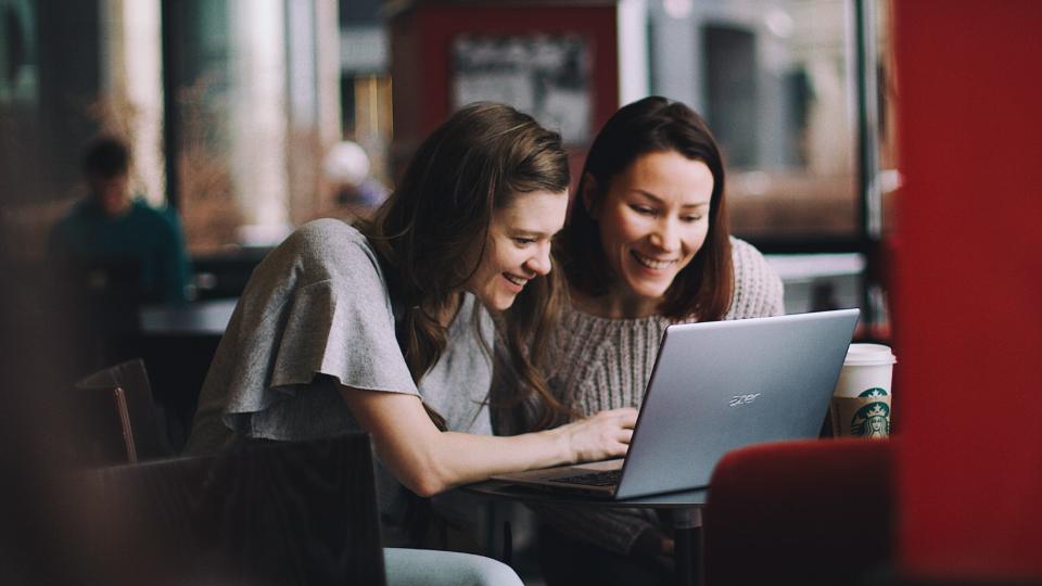Dos mujeres trabajando en su laptop y riendo sentadas en un café, revisando su cuenta de ads